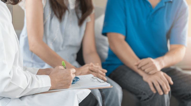 Two people seated with a doctor who is writing notes on a clipboard.