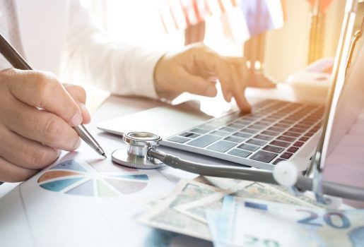 A doctor's hands seen working on a laptop next to a stack of money and a stethoscope. 