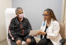 Elexis Williams showing a patient an information packet. The patient wears a black jacket and a face mask.