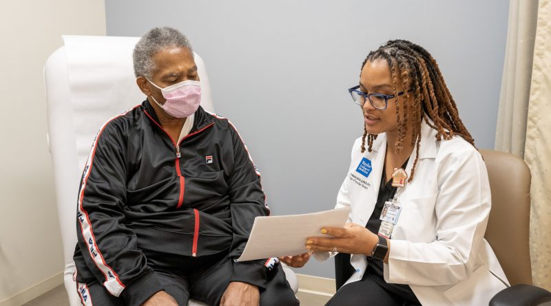 Elexis Williams showing a patient an information packet. The patient wears a black jacket and a face mask.