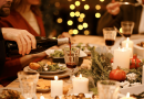 A cheerful holiday spread. Christmas lights in the background illuminate a table where a man pours wine while others eat.