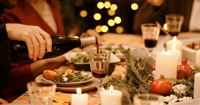 A cheerful holiday spread. Christmas lights in the background illuminate a table where a man pours wine while others eat.