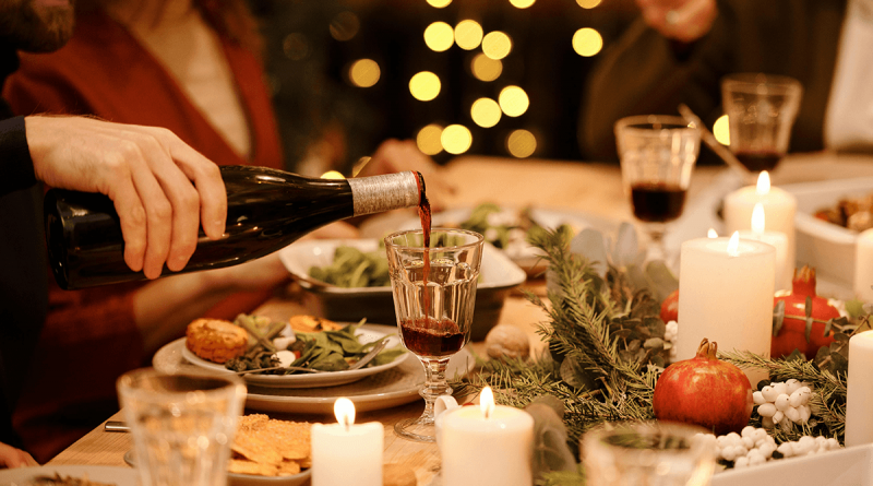 A cheerful holiday spread. Christmas lights in the background illuminate a table where a man pours wine while others eat.