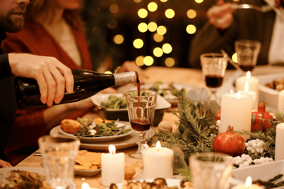 A cheerful holiday spread. Christmas lights in the background illuminate a table where a man pours wine while others eat.