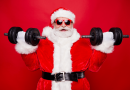 Santa Claus wearing red-rimmed glasses and lifting weights. He has an excited expression as he curls dumbells.