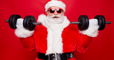 Santa Claus wearing red-rimmed glasses and lifting weights. He has an excited expression as he curls dumbells.