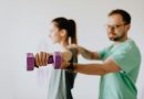 A trainer standing behind a person who is lifting a weight. The lifter holds a purple dumbbell stretched straight to their side, while the trainer guides their arm.