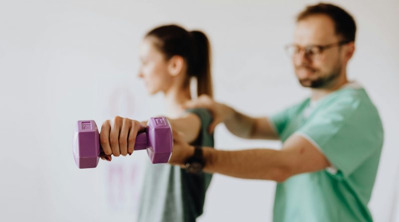 A trainer standing behind a person who is lifting a weight. The lifter holds a purple dumbbell stretched straight to their side, while the trainer guides their arm.