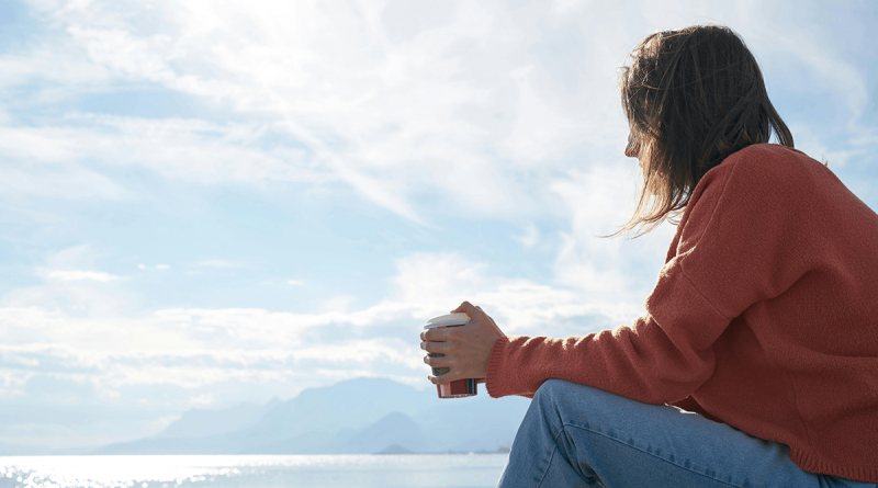 A person in a red sweater holding a cup of coffee while sitting by the ocean, looking off to the horizon.