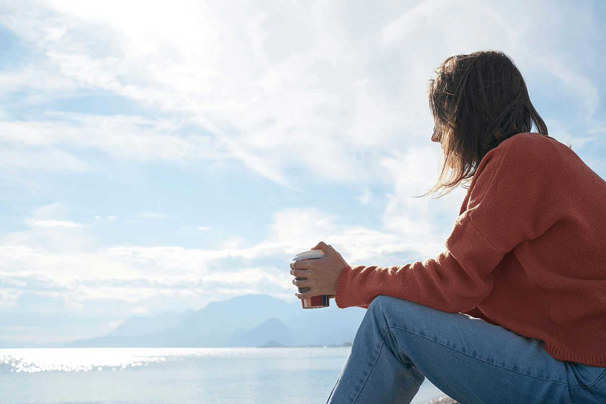 A person in a red sweater holding a cup of coffee while sitting by the ocean, looking off to the horizon.