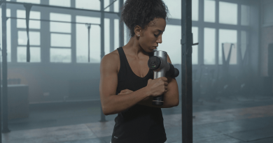 A person using a massage gun on their upper arm. They're dressed in workout gear and seen in a large empty warehouse.