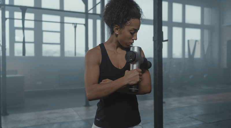 A person using a massage gun on their upper arm. They're dressed in workout gear and seen in a large empty warehouse.