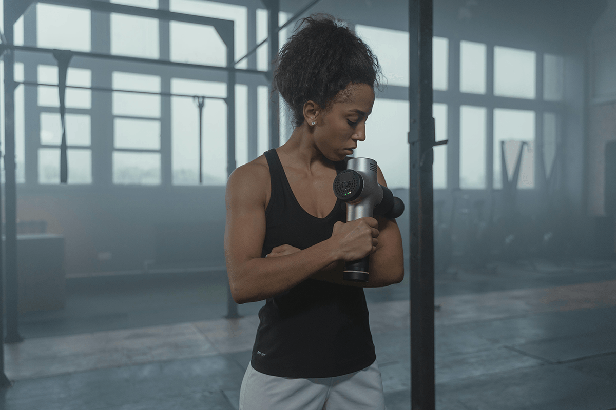 A person using a massage gun on their upper arm. They're dressed in workout gear and seen in a large empty warehouse.