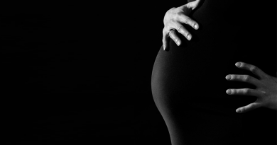 A pregnant person holding their stomach. The photo is in black and white, and the person is wearing a black dress so that their hands are the only part of them standing out.