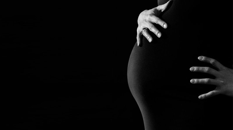 A pregnant person holding their stomach. The photo is in black and white, and the person is wearing a black dress so that their hands are the only part of them standing out.