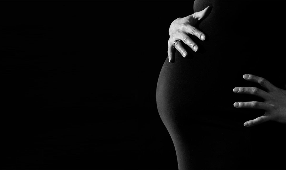 A pregnant person holding their stomach. The photo is in black and white, and the person is wearing a black dress so that their hands are the only part of them standing out.