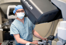 Dr. Kenneth Liao, masked and wearing surgical scrubs, standing behind a large robotic surgery machine.