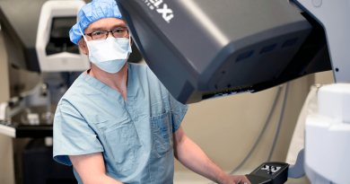 Dr. Kenneth Liao, masked and wearing surgical scrubs, standing behind a large robotic surgery machine.