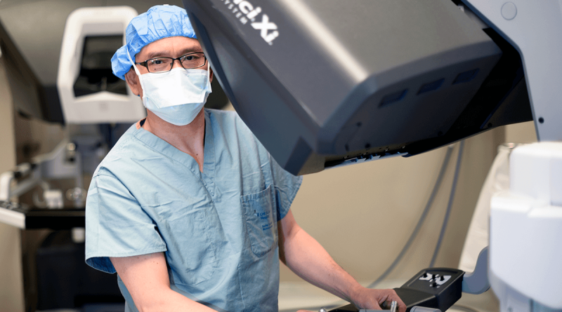 Dr. Kenneth Liao, masked and wearing surgical scrubs, standing behind a large robotic surgery machine.