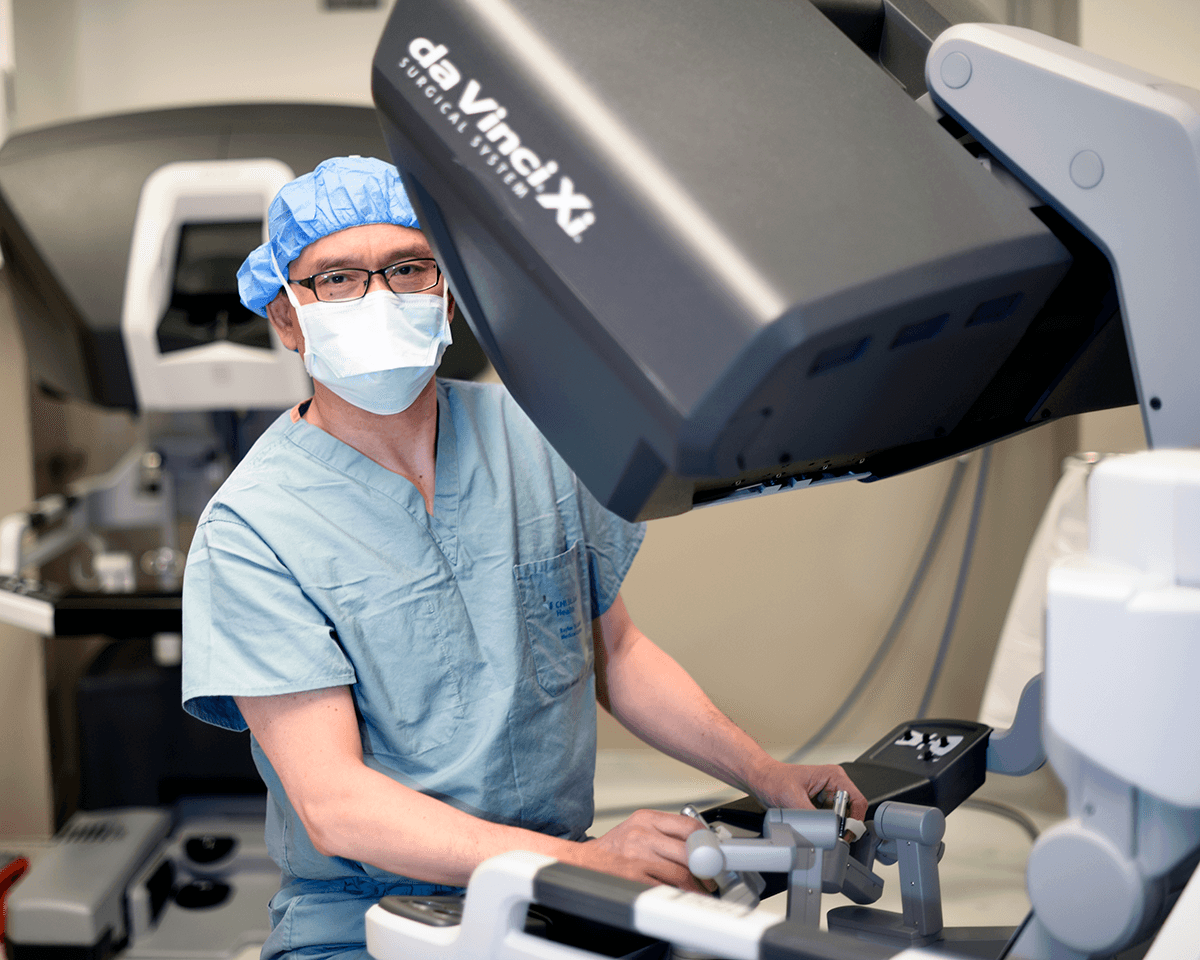 Dr. Kenneth Liao, masked and wearing surgical scrubs, standing behind a large robotic surgery machine. 