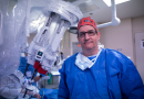 Dr. Richard Link wearing blue scrubs and a bandana on his head stand in front of a robotic surgery system.