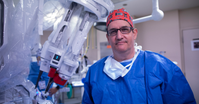 Dr. Richard Link wearing blue scrubs and a bandana on his head stand in front of a robotic surgery system.