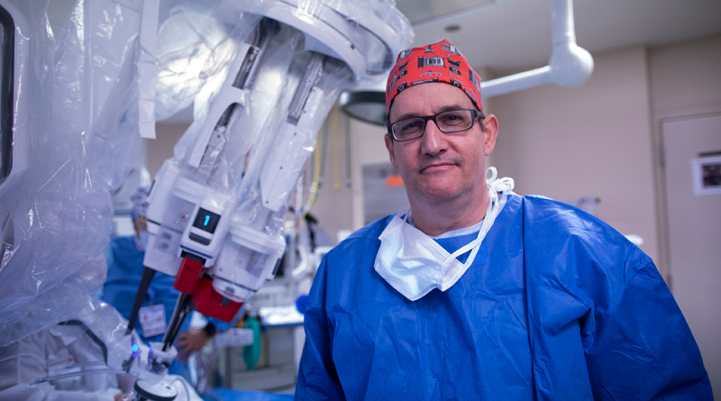 Dr. Richard Link wearing blue scrubs and a bandana on his head stand in front of a robotic surgery system.