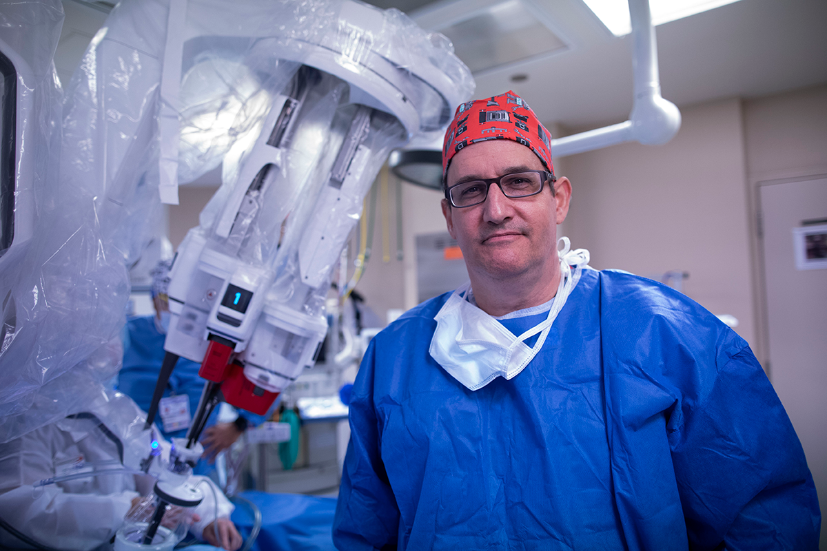 Dr. Richard Link wearing blue scrubs and a bandana on his head stand in front of a robotic surgery system.