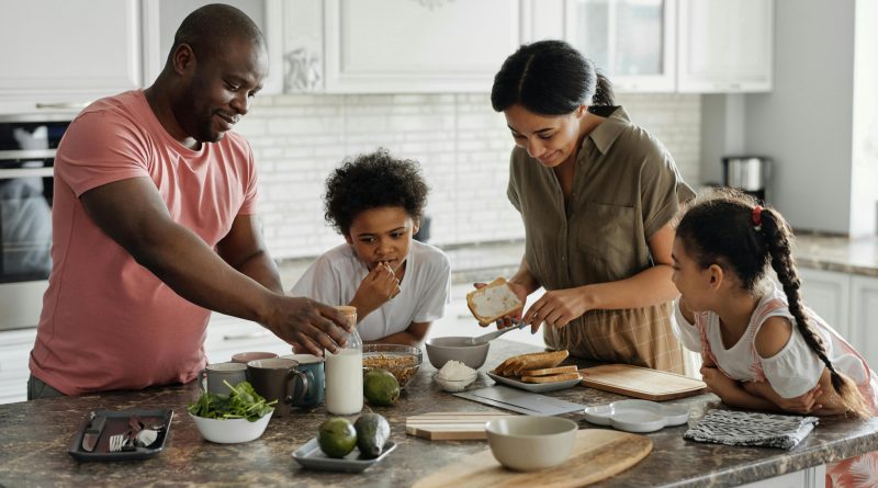 A family in the kitchen putting together a meal, with bowls of cereal, avocados and slices of toast.