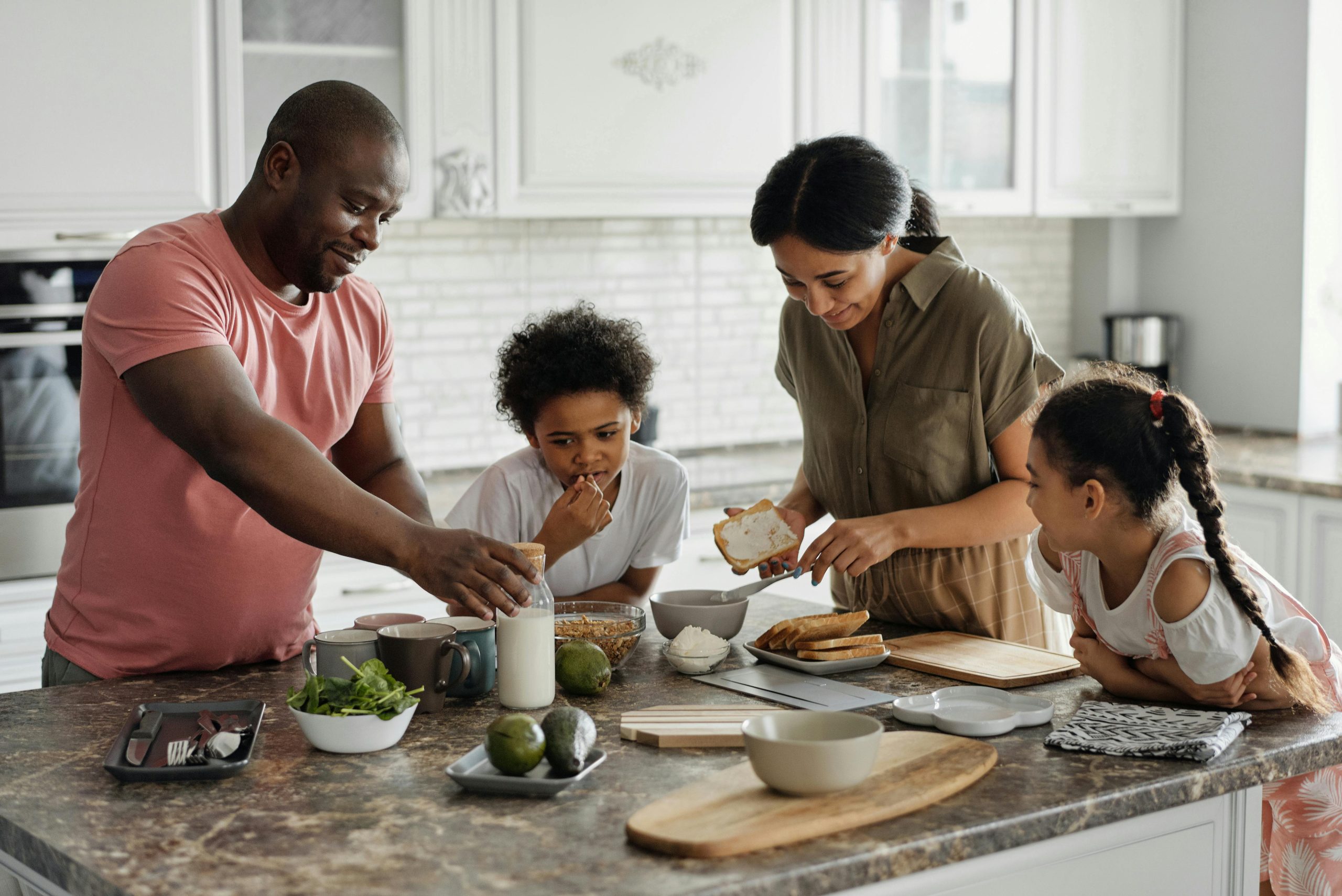 A family in the kitchen putting together a meal, with bowls of cereal, avocados and slices of toast.