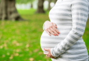 A pregnant person's torso seen from the side, their arms wrapped around their stomach. A lush green wooded area is seen behind them.