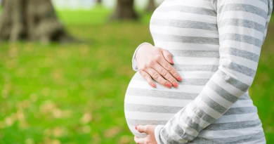 A pregnant person's torso seen from the side, their arms wrapped around their stomach. A lush green wooded area is seen behind them.