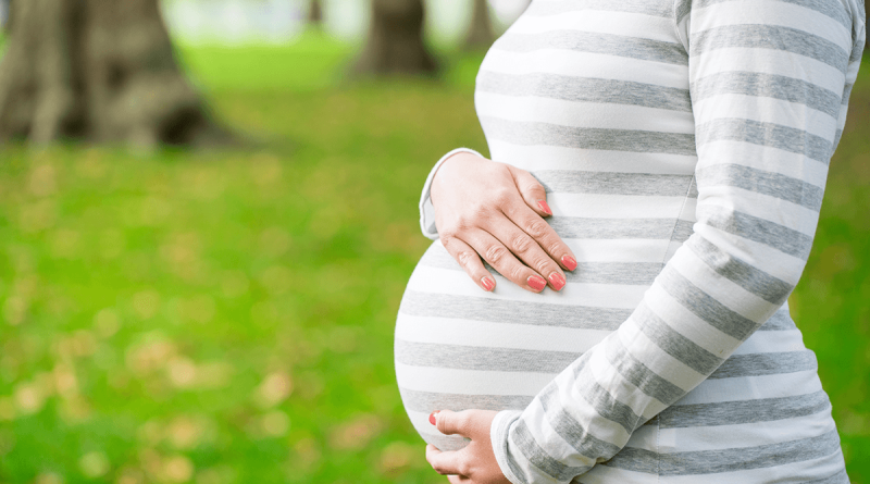 A pregnant person's torso seen from the side, their arms wrapped around their stomach. A lush green wooded area is seen behind them.