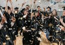 A group of graduates wearing black caps and gowns are celebrating indoors. They are holding diplomas and throwing confetti in the air. The background includes a basketball hoop, indicating that the celebration is taking place in a gymnasium.