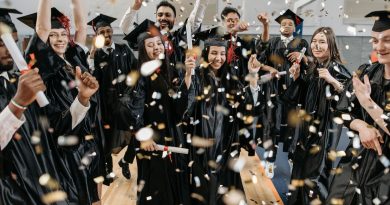 A group of graduates wearing black caps and gowns are celebrating indoors. They are holding diplomas and throwing confetti in the air. The background includes a basketball hoop, indicating that the celebration is taking place in a gymnasium.