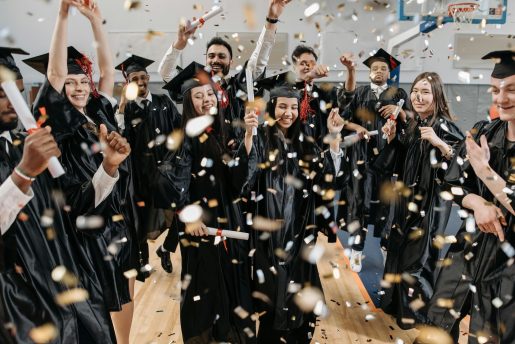 A group of graduates wearing black caps and gowns are celebrating indoors. They are holding diplomas and throwing confetti in the air. The background includes a basketball hoop, indicating that the celebration is taking place in a gymnasium.
