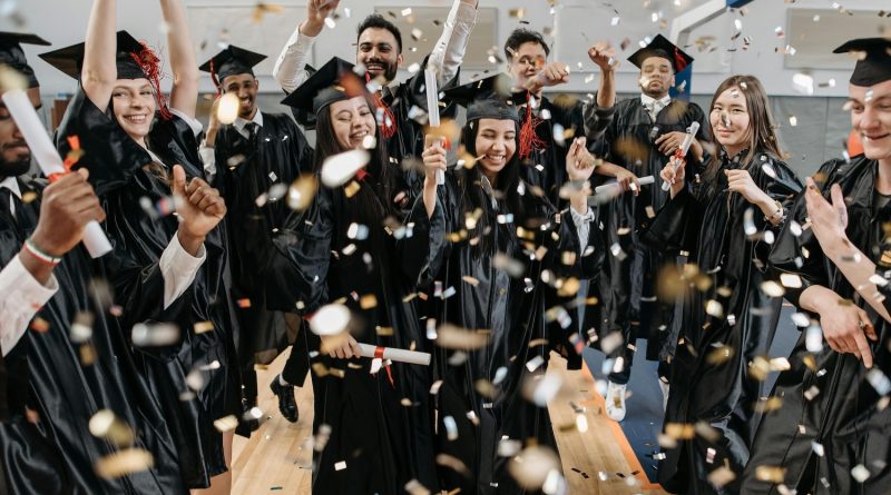 A group of graduates wearing black caps and gowns are celebrating indoors. They are holding diplomas and throwing confetti in the air. The background includes a basketball hoop, indicating that the celebration is taking place in a gymnasium.
