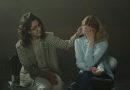 Two individuals are seated in a dimly lit room. The person on the left, dressed in a checkered blazer and light-colored pants, is gently touching the head of the person on the right. The person on the right, wearing a light blue shirt and dark jeans, has their face covered with their hands and appears to be holding a tissue. The scene conveys a moment of comfort and emotional support.