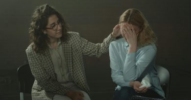 Two individuals are seated in a dimly lit room. The person on the left, dressed in a checkered blazer and light-colored pants, is gently touching the head of the person on the right. The person on the right, wearing a light blue shirt and dark jeans, has their face covered with their hands and appears to be holding a tissue. The scene conveys a moment of comfort and emotional support.