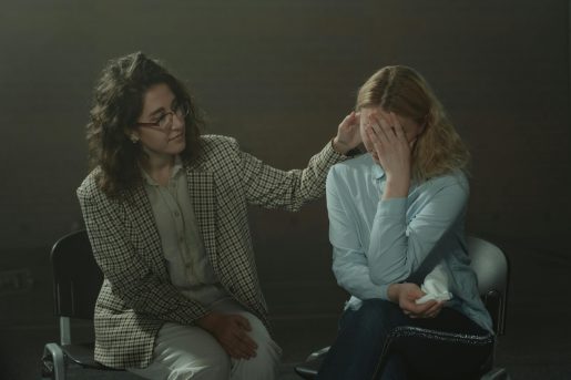 Two individuals are seated in a dimly lit room. The person on the left, dressed in a checkered blazer and light-colored pants, is gently touching the head of the person on the right. The person on the right, wearing a light blue shirt and dark jeans, has their face covered with their hands and appears to be holding a tissue. The scene conveys a moment of comfort and emotional support.