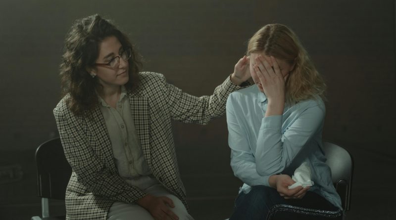 Two individuals are seated in a dimly lit room. The person on the left, dressed in a checkered blazer and light-colored pants, is gently touching the head of the person on the right. The person on the right, wearing a light blue shirt and dark jeans, has their face covered with their hands and appears to be holding a tissue. The scene conveys a moment of comfort and emotional support.