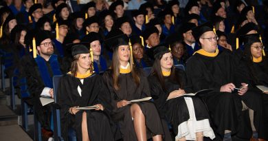 A large group of graduates is seated in an auditorium, wearing black graduation gowns and caps with yellow tassels. Some individuals are holding programs or booklets. The audience behind them is filled with people, some of whom are also dressed in academic regalia.