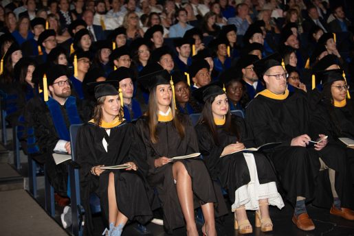 A large group of graduates is seated in an auditorium, wearing black graduation gowns and caps with yellow tassels. Some individuals are holding programs or booklets. The audience behind them is filled with people, some of whom are also dressed in academic regalia.