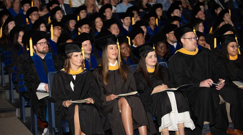 A large group of graduates is seated in an auditorium, wearing black graduation gowns and caps with yellow tassels. Some individuals are holding programs or booklets. The audience behind them is filled with people, some of whom are also dressed in academic regalia.
