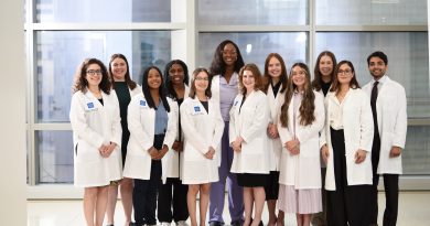A group of twelve people in white coats stand together in a bright room with large windows.