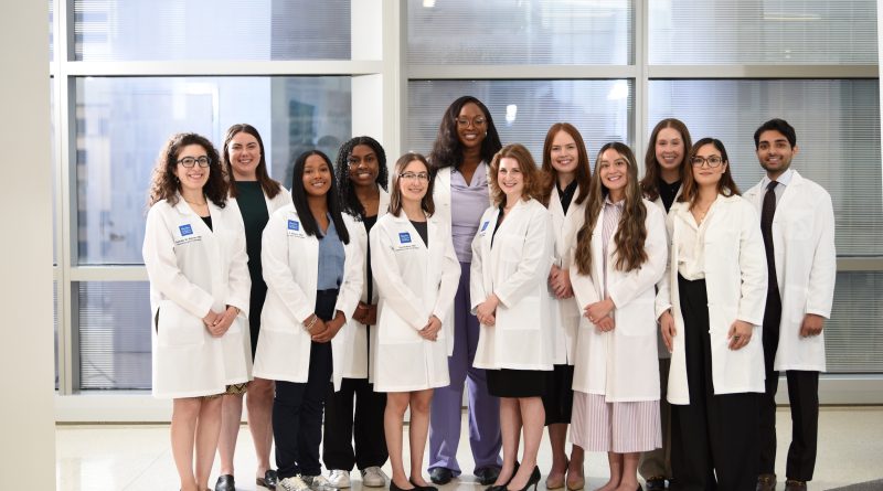 A group of twelve people in white coats stand together in a bright room with large windows.
