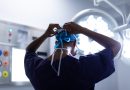 A medical professional, seen from behind, is tying the strings of a blue surgical cap. The individual is wearing a dark scrub top and is in an operating room with bright surgical lights and various medical equipment visible in the background.