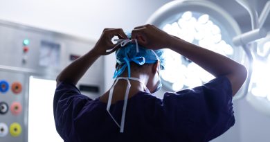 A medical professional, seen from behind, is tying the strings of a blue surgical cap. The individual is wearing a dark scrub top and is in an operating room with bright surgical lights and various medical equipment visible in the background.