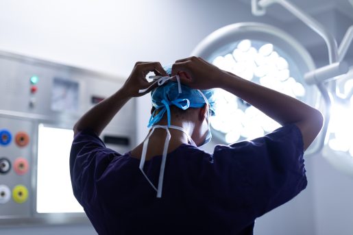 A medical professional, seen from behind, is tying the strings of a blue surgical cap. The individual is wearing a dark scrub top and is in an operating room with bright surgical lights and various medical equipment visible in the background.