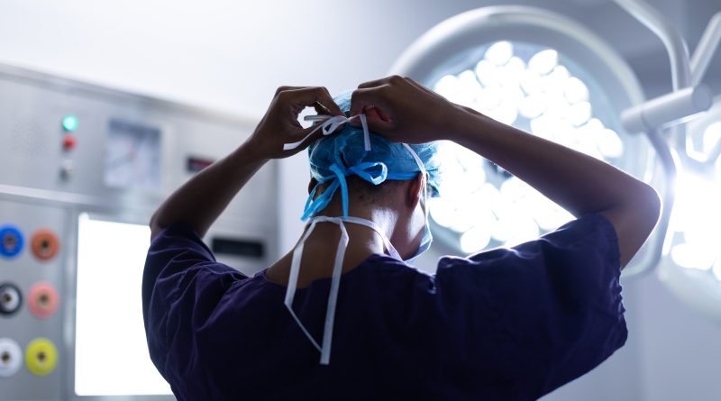 A medical professional, seen from behind, is tying the strings of a blue surgical cap. The individual is wearing a dark scrub top and is in an operating room with bright surgical lights and various medical equipment visible in the background.